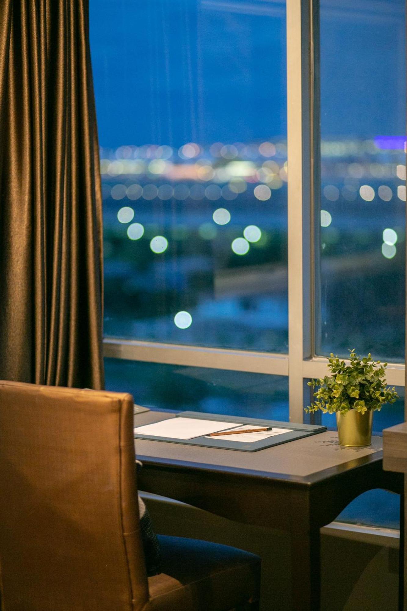 סאמוט פראקאן Siam Mandarina Hotel - Free Bkk Airport Shuttle מראה חיצוני תמונה The photo depicts an indoor workspace with a view of a cityscape in the background. There is a stylish desk accompanied by a comfortable chair. On the desk, there are some blank sheets of paper resting on a blue folder or mat. In the foreground, ther